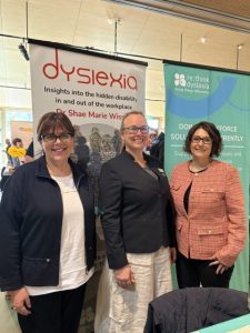 The image features three women standing together at an event with two banners behind them. The banners focus on dyslexia awareness, with one highlighting insights into dyslexia in the workplace and the other promoting alternative approaches to supporting individuals with dyslexia. The women are smiling and dressed in professional attire.